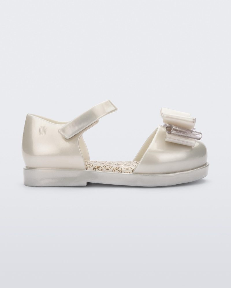 Side view of a white metallic Mini Melissa sandal with a Barbie bow detail on the front toe, white metallic ankle strap and a Barbie logo sole