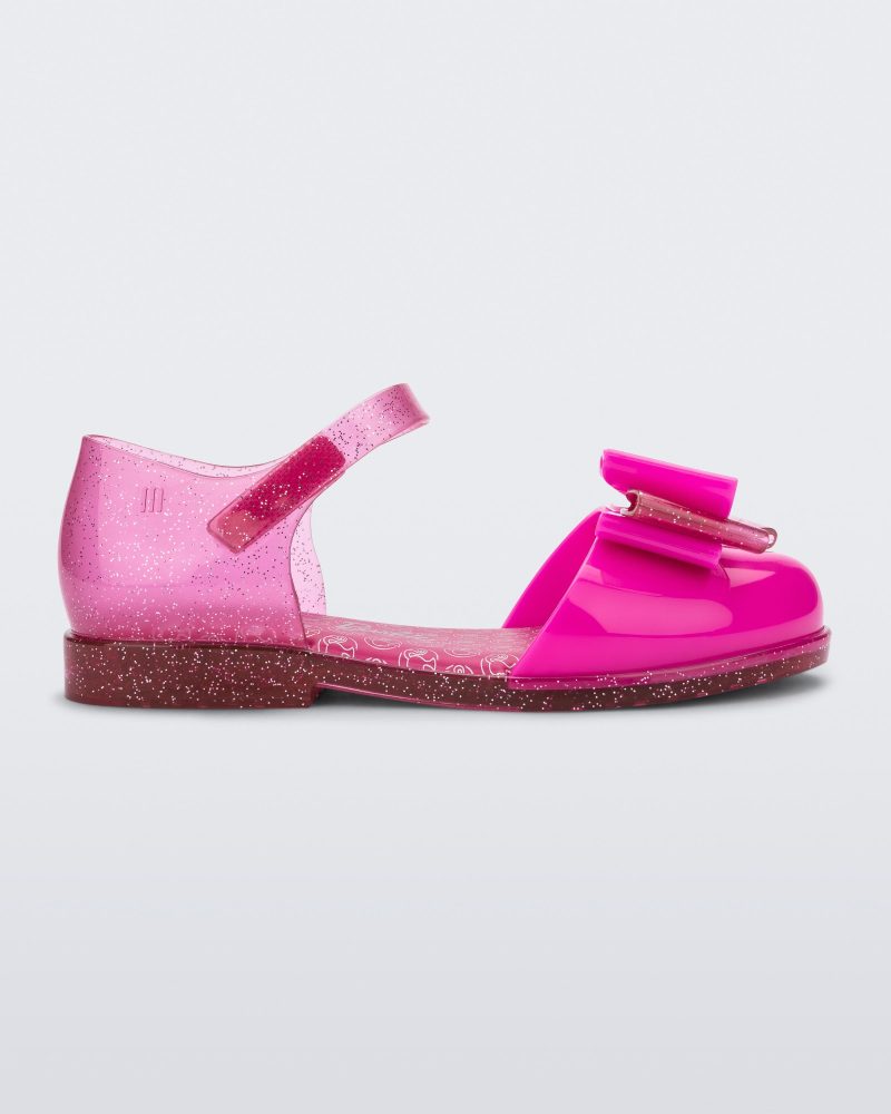 Side view of a glitter pink Mini Melissa sandal with a Barbie bow detail on the front toe, pink glitter ankle strap and a Barbie logo sole