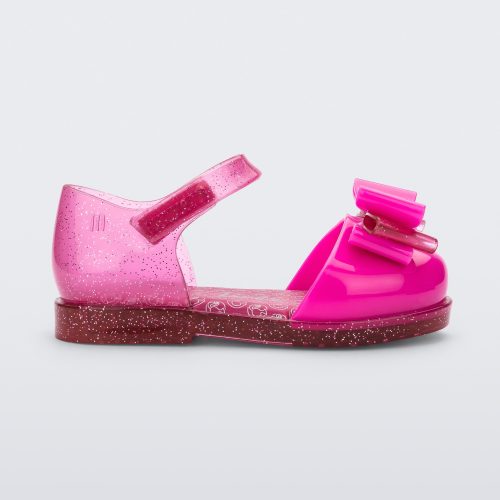 Side view of a glitter pink Mini Melissa sandal with a Barbie bow detail on the front toe, pink glitter ankle strap and a Barbie logo sole