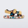 Side view of a beige/mustard Mini Melissa Ping Pong sandal with a black multicolored patterned base, white multicolored sole and yellow velcro straps.