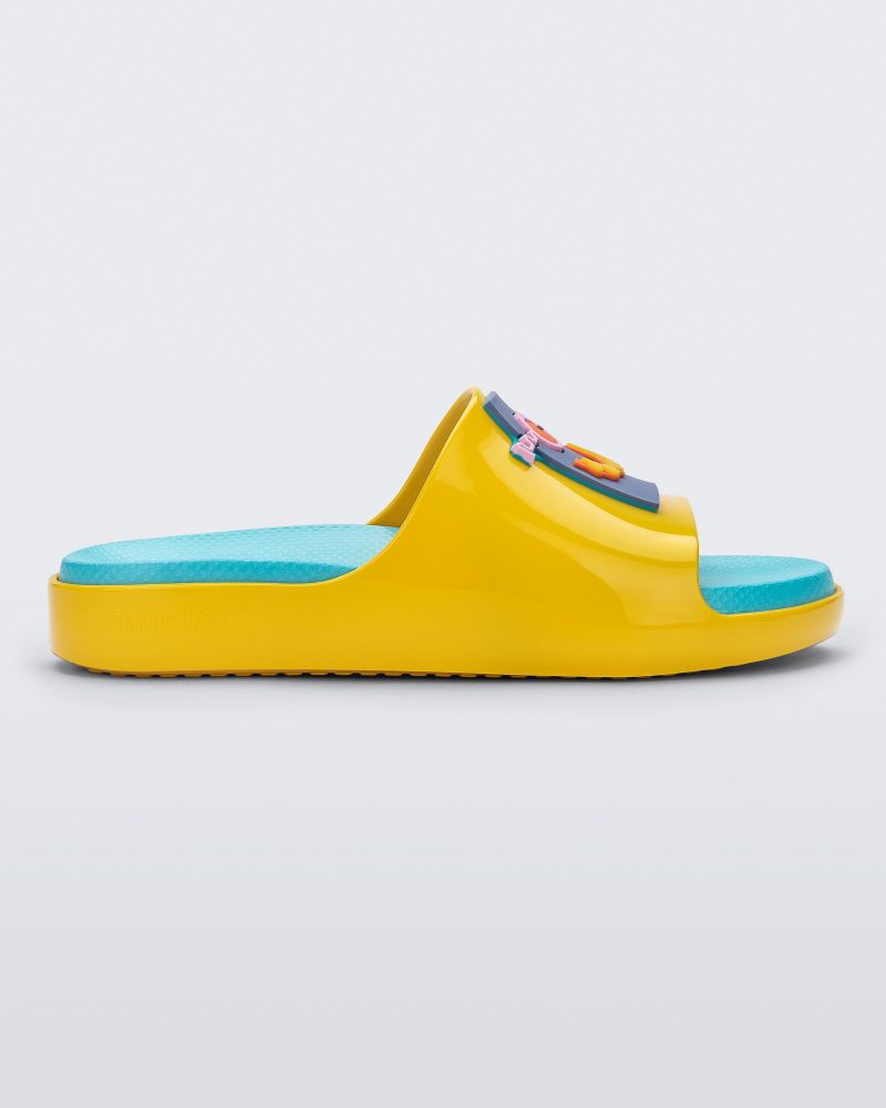 Side view of a pair of yellow Mini Melissa Cloud slide with a blue insole, yellow base and a cartoon drawn smiley face on the front.