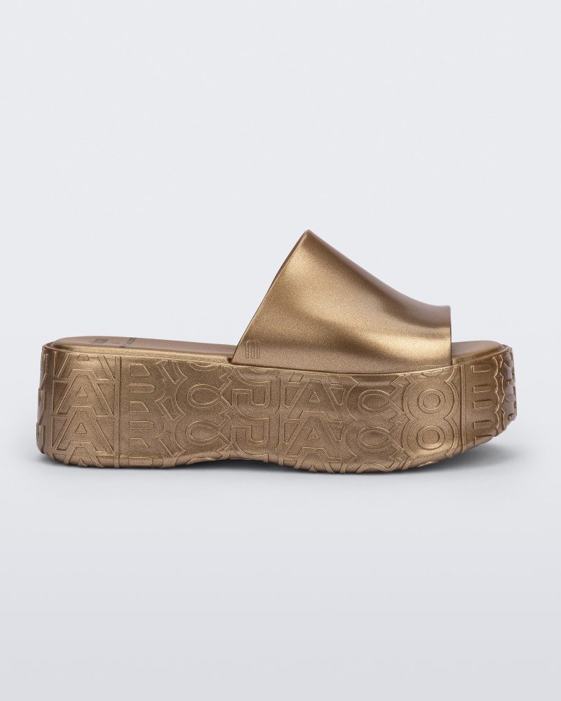 Side view of a gold Becky + Marc Jacobs platform slide.