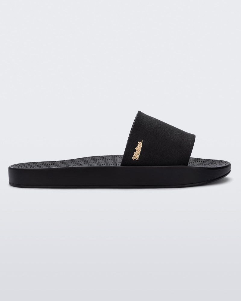 Side view of a Melissa Sun Sunset slide sandal in black with gold Melissa logo on the top side corner of the strap.