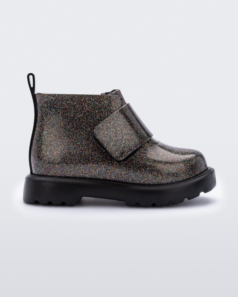 An outter side view of a black/glitter multicolor Mini Melissa Chelsea boot with a multicolor glitter base, velcro front strap and a black sole.